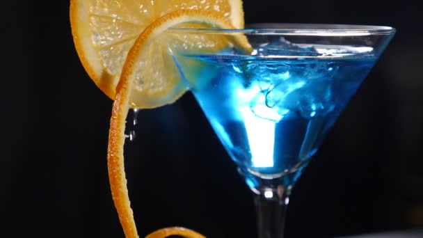 Wunderschönes Getränkevideo in Zeitlupe. Nahaufnahme einer Orangenscheibe auf blauem Cocktail auf schwarzem Hintergrund, Wassertropfen fallen. Früchte geben Frische und Saftfüllung ab. Konzept — Stockvideo