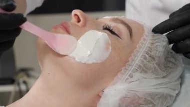Güzellik salonunda. Kozmetik uzmanı kaplıca salonunda kadın yüz derisine maske takıyor. Güzellik uzmanı sopa kullanarak kaldırma kremi sürerken güzel bir kadın spatulayla güzellik prosedürü uyguluyor.