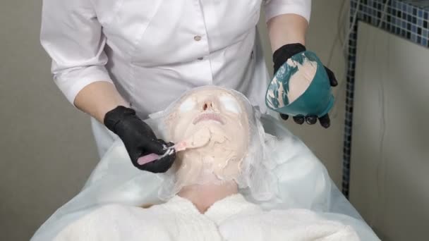 Close-up shot of young woman getting facial treatment in beauty salon. Female client in beauty clinic. beautician in black gloves applying with spatula clay mask on face covered with gauze. 4 k video — Wideo stockowe