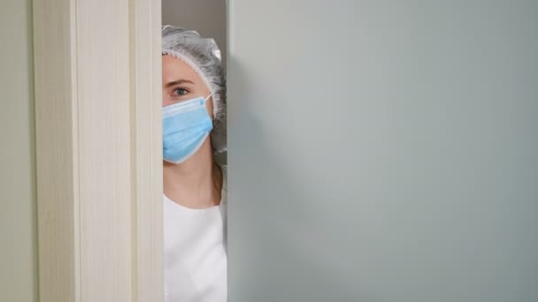 年轻的女医生或护士戴着面罩，打开地面玻璃门，目不转睛地看着相机，而不站在半开着的门或门阶上。临床医生正在检查病房。4 k镜头 — 图库视频影像