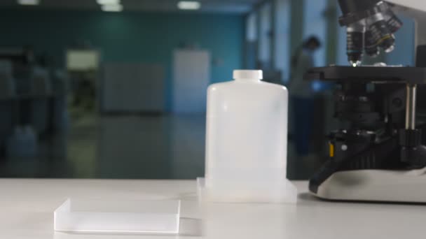 Konsep laboratorium modern. Ilmuwan sarung tangan hitam meletakkan wadah botol plastik putih dengan mikroskop di latar belakang. Salin ruang untuk catatan. 4 K rekaman — Stok Video