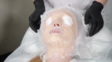 Kozmetik prosedürü. Güzellik uzmanı kadın hasta yüzünden sertleştirilmiş maskeyi çıkarıyor. Yüzü maske altında hareket ediyor. Spa ve güzellik salonu. Genç bir kadın aljinat maskesiyle cilt bakımı yaptırıyor. 4k video
