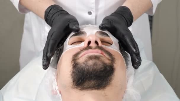 ハンサムな男性のためのスパ療法顔マスクを取得します。4k映像だ。現代のスパサロンで美容師のレセプション。男性の顔に化粧品業者がマスクを保湿. — ストック動画