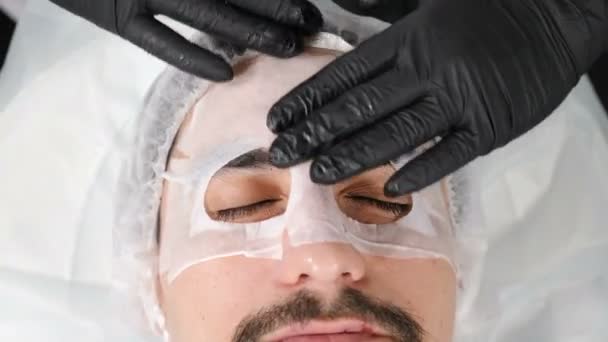 Terapia de spa para hombres guapos con mascarilla facial. Imágenes de 4k. Recepción de un cosmetólogo en el moderno salón de spa. cosmético poner en la cara masculina mascarilla hidratante . — Vídeos de Stock