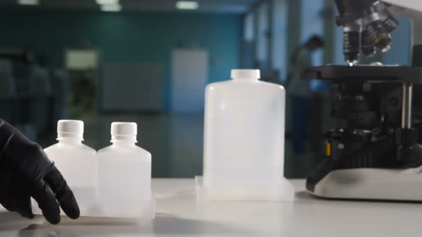 Modern laboratuvar konsepti. Siyah eldivenli bilim adamı arka planda mikroskop olan beyaz plastik şişe kapları koyuyor. Boşluğu nota kopyala. 4k görüntü — Stok video