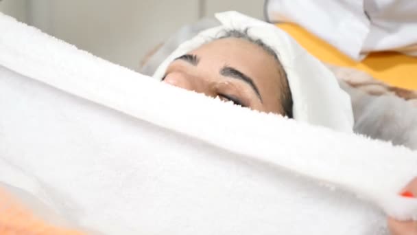 Na clínica de beleza. Esteticista ativando máscara facial com toalha branca quente. Close-up. Jovem mulher bonita recebendo procedimento no salão. rosto feminino sendo envolto por toalha para aquecer a pele facial. Facial — Vídeo de Stock