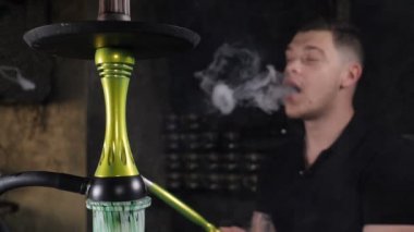 Nargile içen yakışıklı genç adam. Kafkasyalı adam aromalı tütün içiyor. Karanlık arka planda yakın plan duman üfle. Buharın yuvarlak formu havada uçuyor. Sihirli sanat sisi, kötü alışkanlık konsepti. yavaş