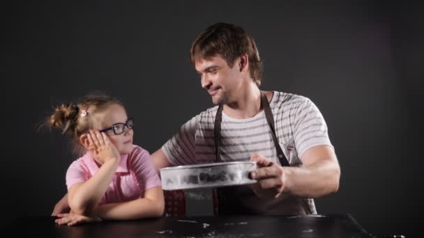 Profissional padeiro masculino e sua linda filha peneirando farinha através da peneira na mesa sobre fundo preto. Cozinha tradicional deliciosa Pizza italiana. Partículas brancas a cair. Movimento lento. hd completo — Vídeo de Stock