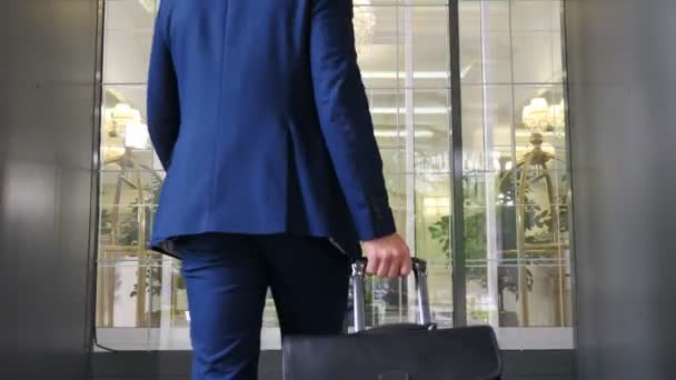 Rear view of young businessman wearing dark suit entering hotel going thrioug glass doors. Man carrying case luggage and briefcase. Tourist concept. 4k footage — Stock Video