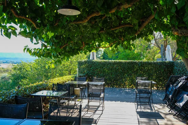 Glass tables on deserted wooden terrace under magnificent tree. Deep sunshine. Provence tourism. Joucas.