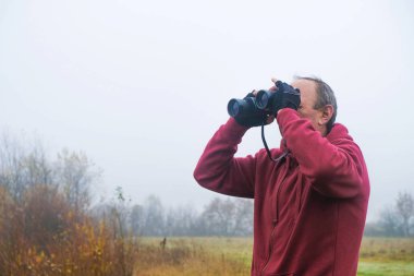59 yaşında kırmızı elbiseli bir adam dürbünle bakar. Sonbahar sisli günü. Uzayı kopyala Kişisel izolasyon kavramı.