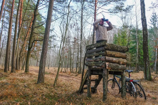 A 59 year old man watches binoculars. Bicycle stands beside a wooden observation tower. Hunting grounds in Ukraine. Self-isolation concept.