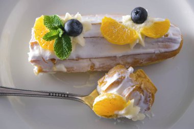 Kremalı kek ve beyaz tabakta taze meyve. Yabanmersini, mandalina dilimleri ve nane yaprakları. Kaşık içinde çocuk oyuncağı..