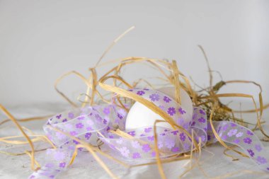 Beyaz arka planda saman ve bayramlık Paskalya kurdelesi olan doğal beyaz yumurta. Mor çiçekli kurdele. Boşluğu kopyala.