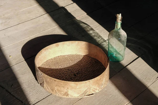 Rural still life with an empty bottle, old sieve and geometric shadows on a rough wooden surface. Bottle in cobweb and dust closed with paper cork.