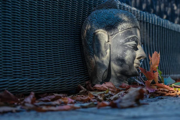 Smiling Buddha statue head in profile on wicker surface near red leaves in morning sun. Mass produced statues. Provence tourism. Changing the philosophy of life concept.