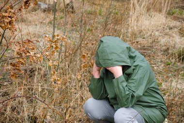 Felakette bir endişe ve umutsuzluk sahnesi. Koruyucu kapüşonlu elbiseli yalnız bir kadın başını eğerek oturdu. Etrafında kuru bitkiler var..
