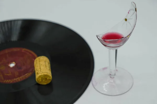 Broken vintage wine glass with red wine and an old vinyl record. On a vinyl record is a wine cork. Still life in art deco style.
