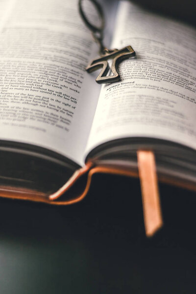 A brown leather bible is opened and set down on a table. In the middle of the open center binding rests a hollow rustic looking cross pendant. The pendent is set in the center of the binding. 