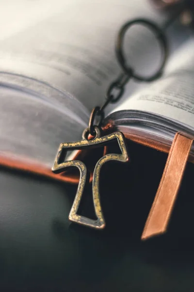 A brown leather bible is opened and placed on a black reflective table top. A rustic metallic cross is hanging out of the bible from the center binding of the open book. The cross pendent slightly rests on the table top.