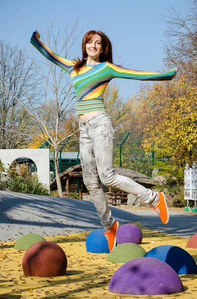 Glad rödhårig ung söt flicka i ljusa nära gärna hoppar på en färgad lekplats i parken, under solsken — Stockfoto