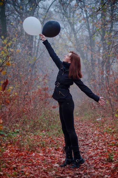 Girl in dark clothing in autumn fog forest, with flying two balloons, black and white. Concept of choice, good and evil