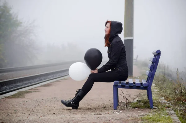 Lonely girl sit on bench on railway platform in fog. She holds black and white balloon. Concept of choice, good and evil