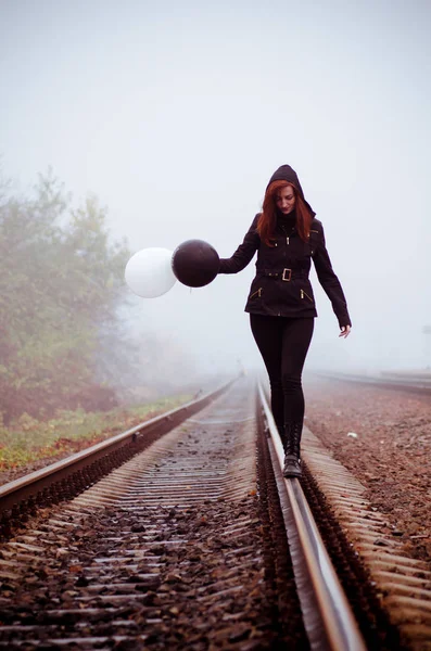 Lonely girl in black clothing stands between rails. She holds balloons, it fly away. Concept of choice, way of life