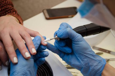 Donanım manikür prosedürü. Mavi eldivenli usta, beyaz kesiciyle tırnaklarındaki jöle cilasını söker.