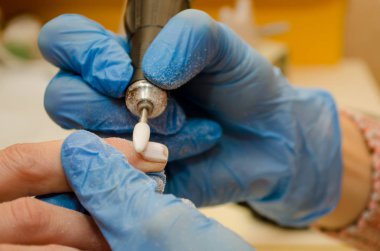 Donanım manikür prosedürü. Mavi eldivenli usta tırnaklarındaki jöle cilasını beyaz keskiyle temizler, toz uçar.