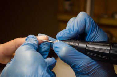 Donanım manikür prosedürü. Mavi eldivenli usta çiviyi uzun bir değirmenle manikür yapar, hizalar ve çivileri temizler.