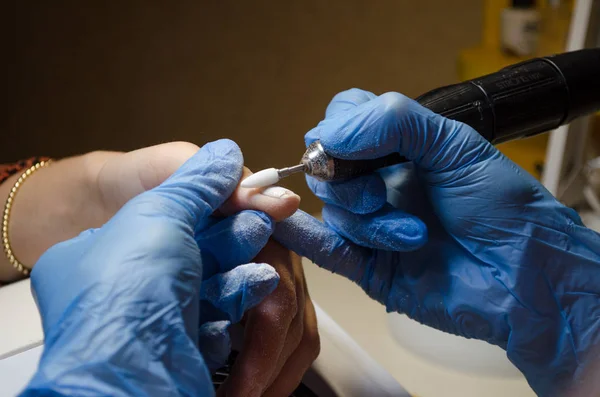 Donanım manikür prosedürü. Mavi eldivenli usta, beyaz kesiciyle tırnaklarındaki jöle cilasını söker.