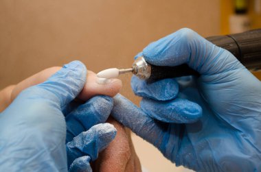 Donanım manikür prosedürü. Mavi eldivenli usta tırnaklarındaki jöle cilasını beyaz keskiyle temizler, toz uçar.