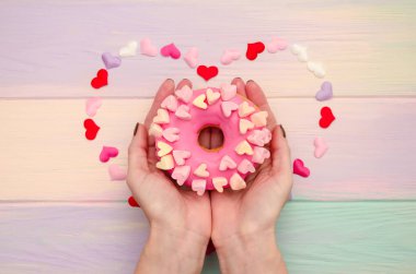 Tatlı pembe donut, kremalı ve dekorasyonlu, kalpler gökkuşağında, ahşap arka planda, kadın ellerinde.