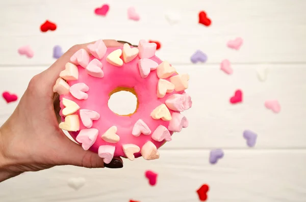 Свиной розовый пончик с сосульками и украшением сердечками на радужном деревянном фоне в женской руке — стоковое фото
