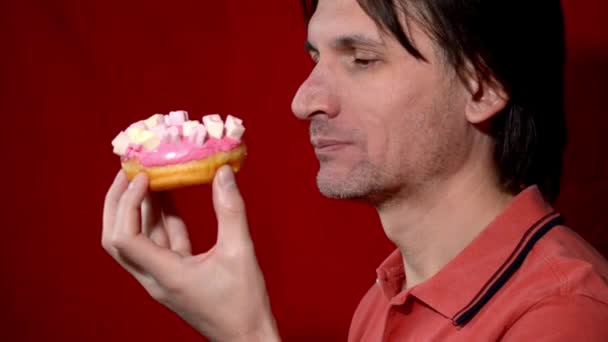 Жорстокий чоловік з темним волоссям у рожевій сорочці їсть рожевий пончик на червоному тлі — стокове відео