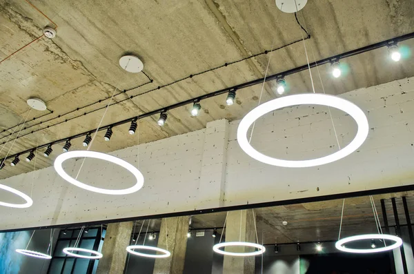 White circular ring shaped lamps hang under concrete ceiling with external wiring and black fixtures, with mirror