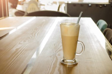 Tall transparent glass beaker with a handle, with foam latte inside and spoon, on wooden table in a cafe in the sunshine