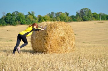Avrupalı genç, kızıl saçlı, ince bir kız siyah ve sarı giysiler içinde samanlığı güneşe doğru itiyor.
