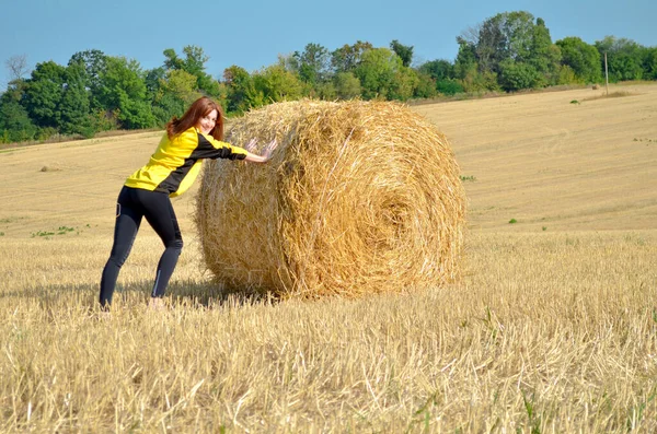 Mladá zrzavá štíhlá dívka evropského vzhledu v černém a žlutém oblečení tlačí kupku sena na pole pod sluncem — Stock fotografie