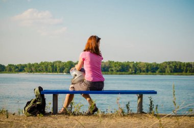 Pembe ve gri elbiseli genç kızıl saçlı kız gün batımında motosiklet kaskıyla nehrin yanındaki bankta oturuyor.