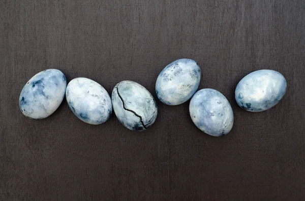 Six blue marble eggs, one of them with a crack, painted in hibiscus tea, lie on a black background with a sprinkle of butterflies
