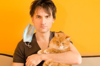 Kırmızı kedili yorgun bir adam mavi maskeyi yüzünden çıkardı, kulaklarına tutundu, karantina tamamlandı, sarı arka plan