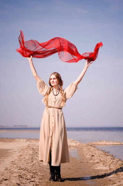 Young beautiful caucasian blond woman, with minimal makeup, in light dress, red scarf against blue sky, water and sand