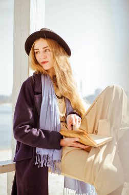 Boho elbisesi, paltosu ve şapkası içinde, güneş ışınları altında cankurtaran evinde kitap okuyup gülümseyen, beyaz tenli genç sarışın kız.
