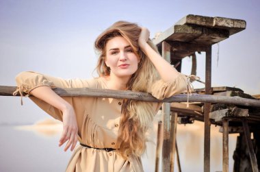 Kafkasyalı güzel sarışın kız, açık renk elbiseli, mavi gökyüzünün arka planında, eski ahşap köprüde oturuyor.