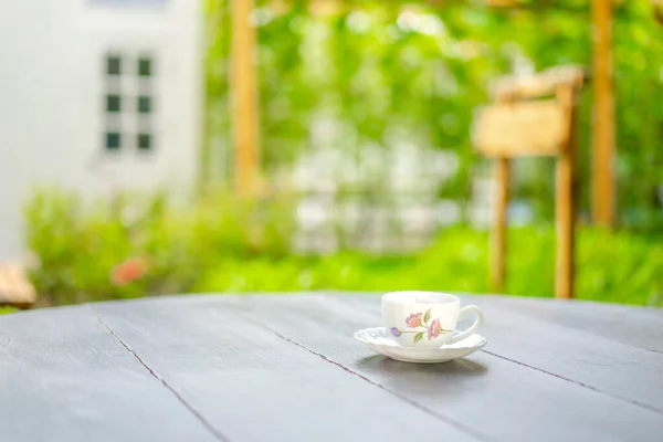 British tea cup style with drawing pink flower pattern on it, on the wood dark brown table in the morning with blur garden and window.