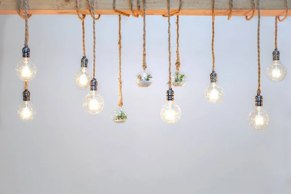 Decorated cozy lamps stick in the rope and little tiny tree with small white rock inside the ball glass hang on to big wood log in front of white background.