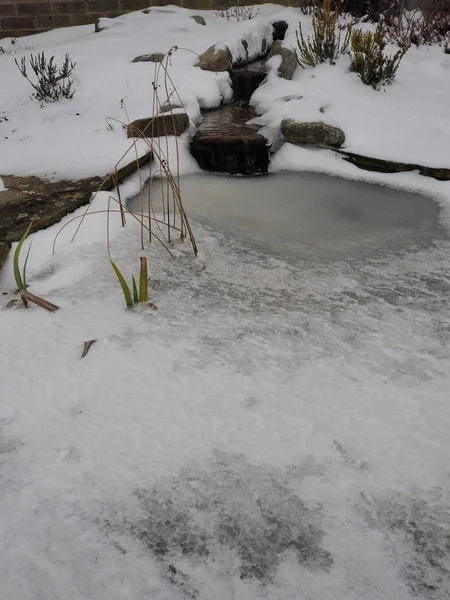 Stagno da giardino con ruscello e cascata con neve e ghiaccio ghiacciato sull'acqua Immagine Stock