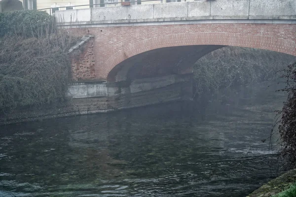 Křivka vodní kanál vedoucí pod mostem přes v mlze, stromy, trávu a towpath — Stock fotografie